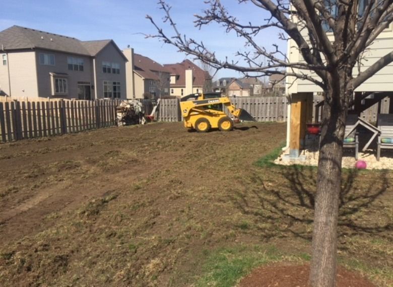 Yellow skid steer leveling soil in a backyard, near a fence and houses.