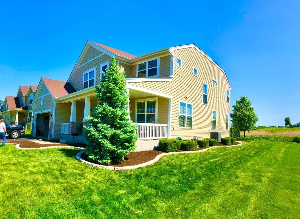 Two-story beige house with green lawn, front porch, and tall evergreen tree under a blue sky.
