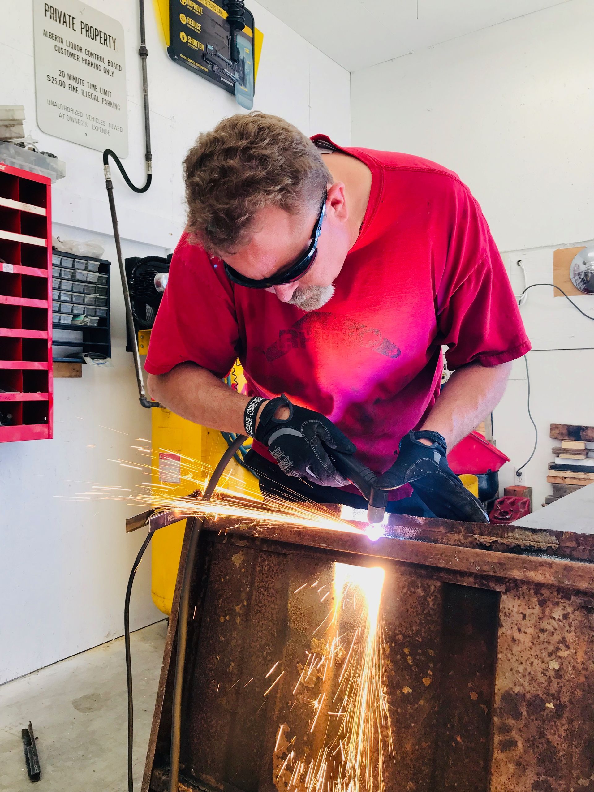 Man in red shirt welds metal in a workshop, sparks flying, wearing safety glasses.