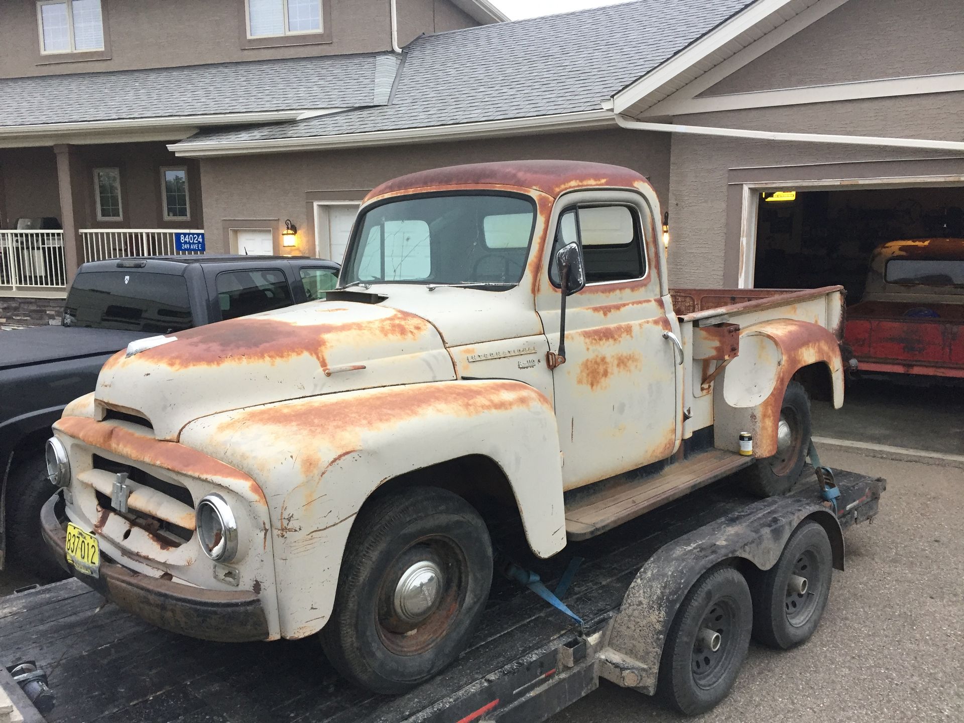 Vintage rusty white pickup truck on a trailer in front of a house and garage.
