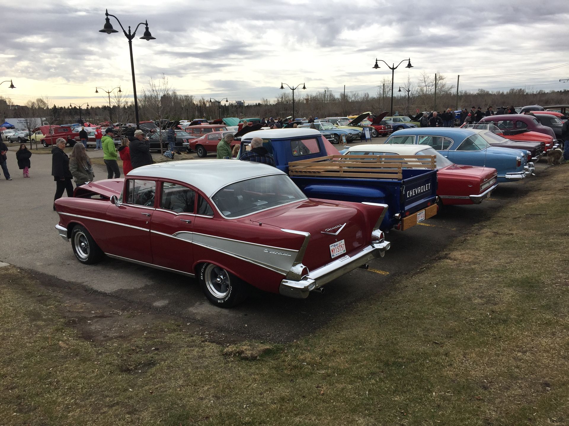 Classic cars parked in a row at an outdoor car show, with people milling about on a cloudy day.