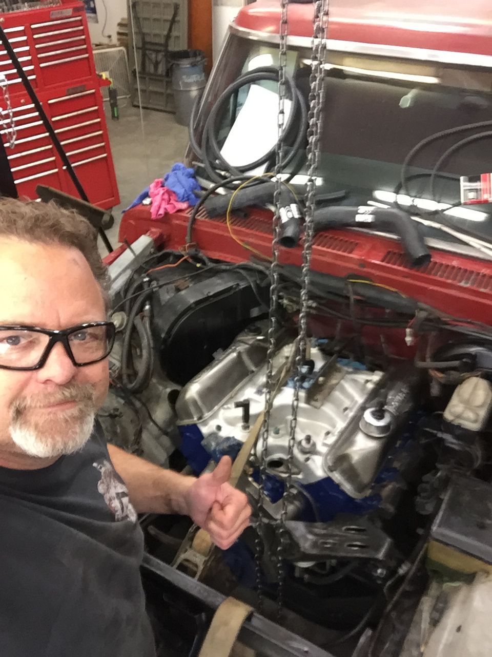 Man gives thumbs-up next to car engine being lowered. Red truck and toolbox are visible.