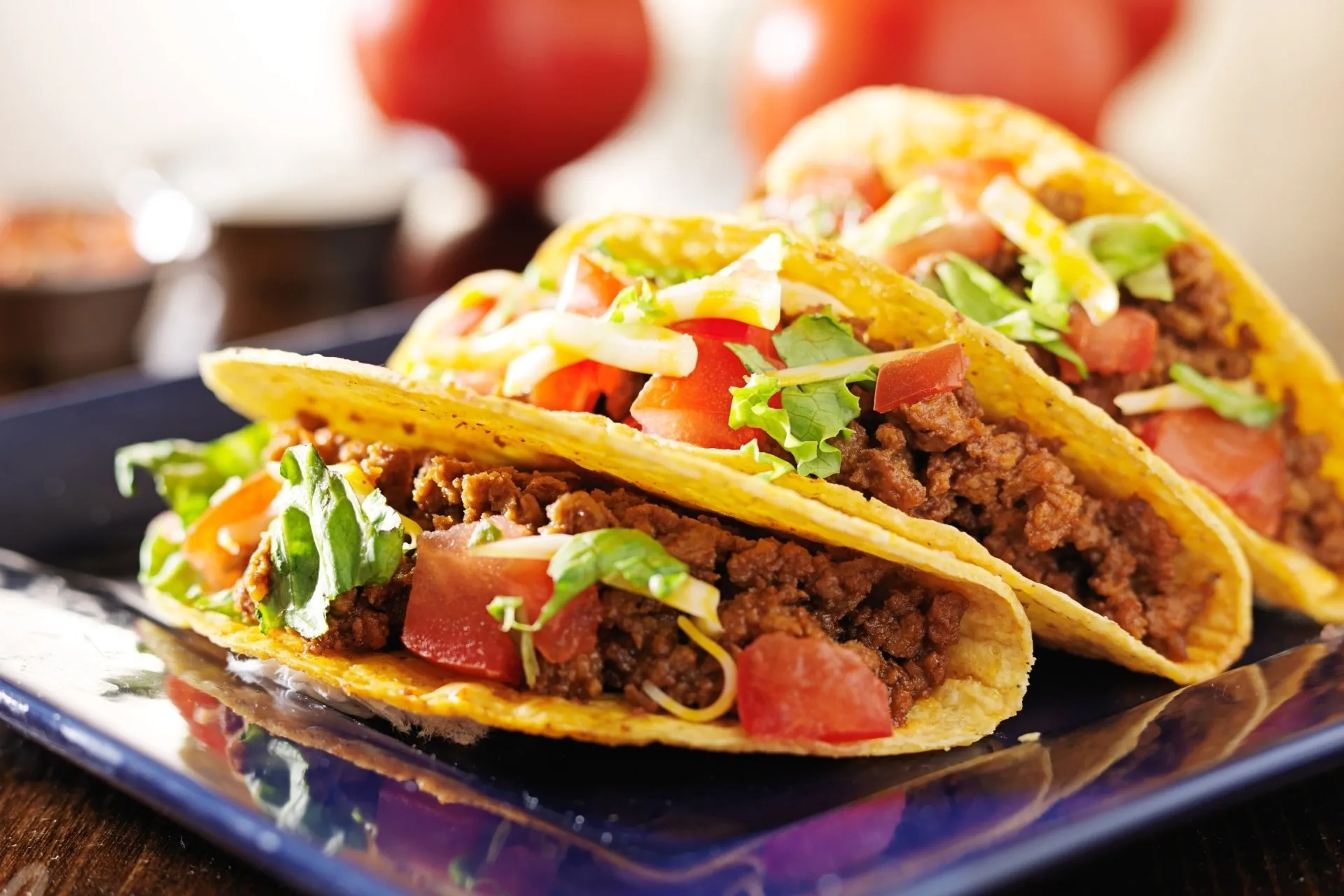 Three loaded tacos on a blue plate, featuring ground meat, tomatoes, lettuce, and cheese.
