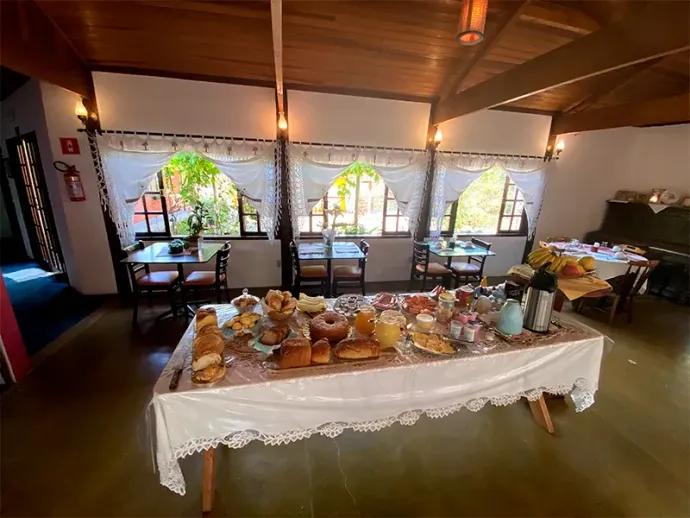 Buffet de café da manhã servido em uma mesa comprida, em uma sala de jantar com janelas e teto com vigas de madeira.