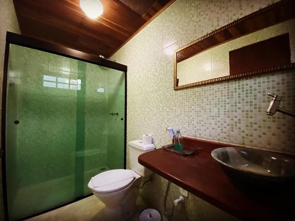 Banheiro com chuveiro em tom esverdeado, vaso sanitário, bancada marrom e paredes revestidas com azulejos em mosaico.