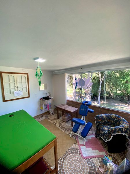 Sala de massagem com maca verde, cadeira de massagem, poltrona e janela com vista para as árvores.