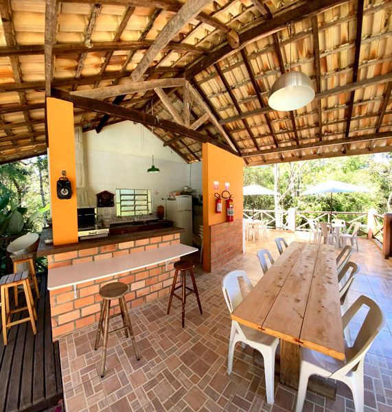 Área de refeições ao ar livre com bar, mesas e cadeiras sob um telhado de telhas. Dia ensolarado.