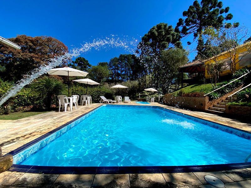 Piscina com fonte, rodeada por árvores, guarda-sóis e mobiliário de jardim num dia ensolarado.
