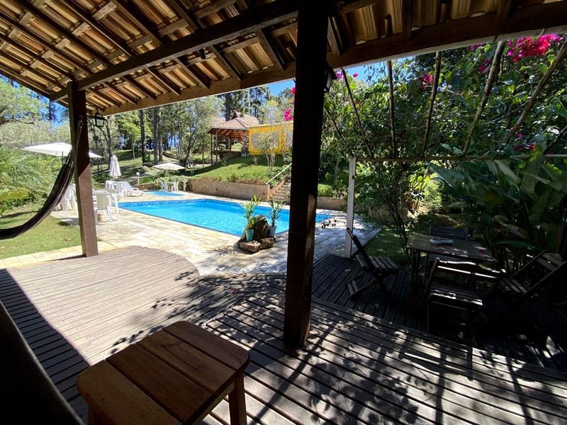 Varanda coberta com vista para a piscina e vegetação exuberante. Mesa e cadeiras de madeira, rede e paisagem ensolarada.