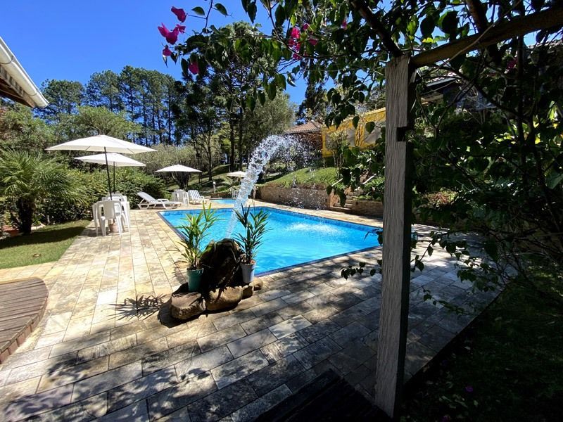 Piscina com fonte, rodeada por um pátio com mesas, cadeiras e vegetação exuberante num dia ensolarado.