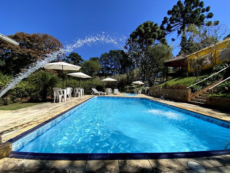 Piscina com fonte e espreguiçadeiras, rodeada por árvores e um céu ensolarado.