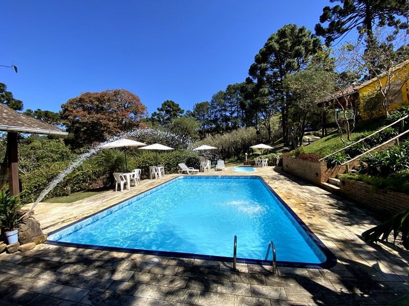 Piscina com fonte, mesas e cadeiras em um dia ensolarado. Árvores ao fundo.