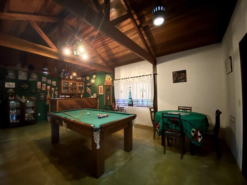 Sala de jogos com mesa de bilhar, mesa de jantar, bar e parede decorativa. Teto de madeira e decoração em tons de verde e branco.