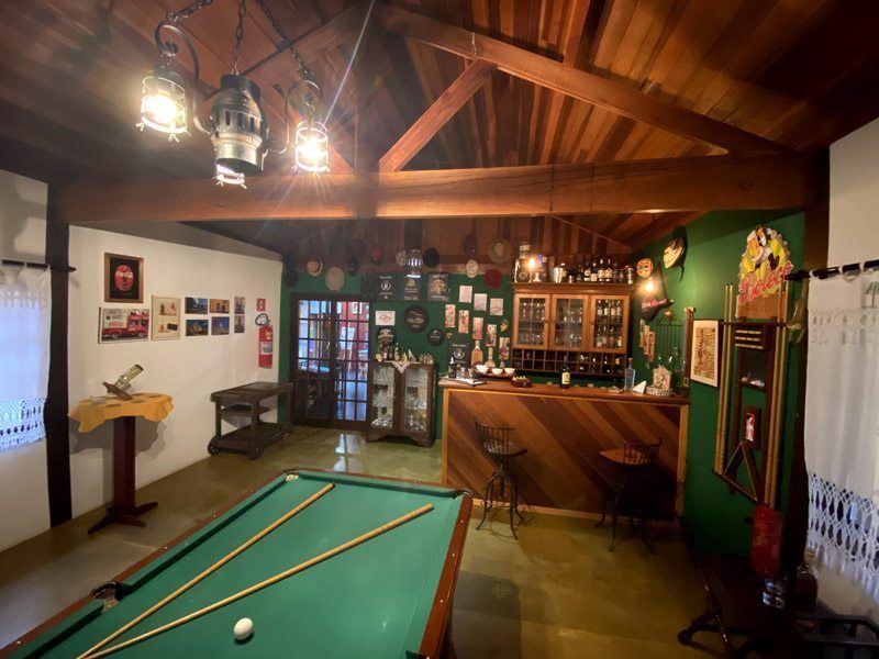 Salão de bilhar com bar, teto de madeira e paredes verdes. Uma mesa de bilhar em primeiro plano com bolas e tacos.