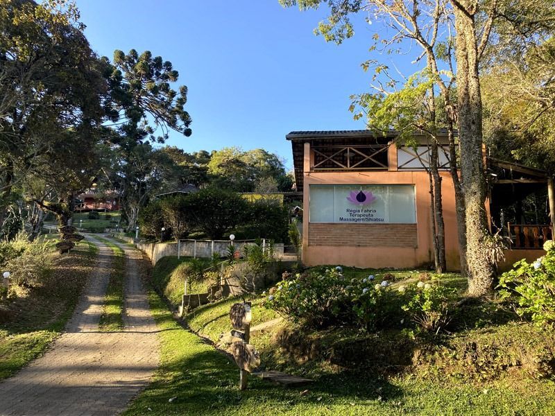 Uma estrada de terra leva a um edifício com uma placa e uma varanda, situado em um ambiente natural e verdejante.