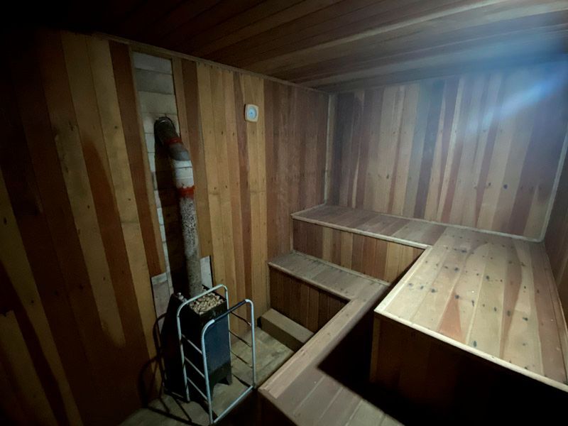 Interior de sauna de madeira com bancos em dois níveis. Um fogão e uma chaminé são visíveis à esquerda.
