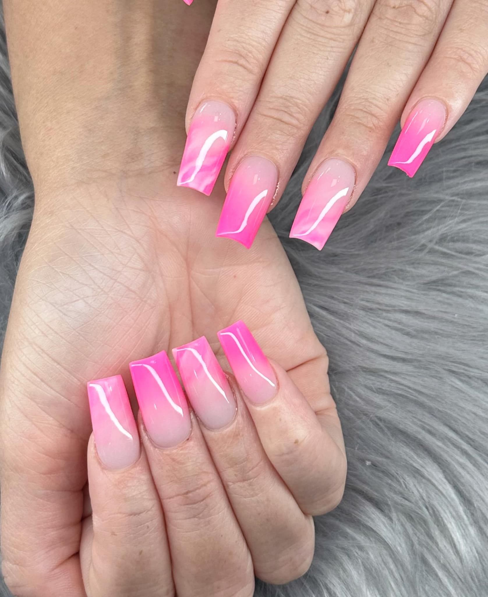 Hands displaying long, square-shaped acrylic nails featuring a pink ombre design against a grey faux-fur background.