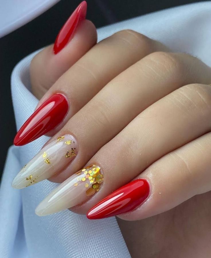 A hand with long, almond-shaped nails featuring a mix of glossy red polish and milky white nails with gold foil accents.