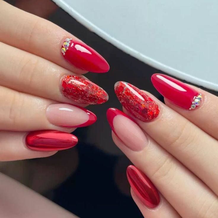 Close-up of hands with almond-shaped nails in a mix of red chrome, red glitter, and red French tip styles with crystals.