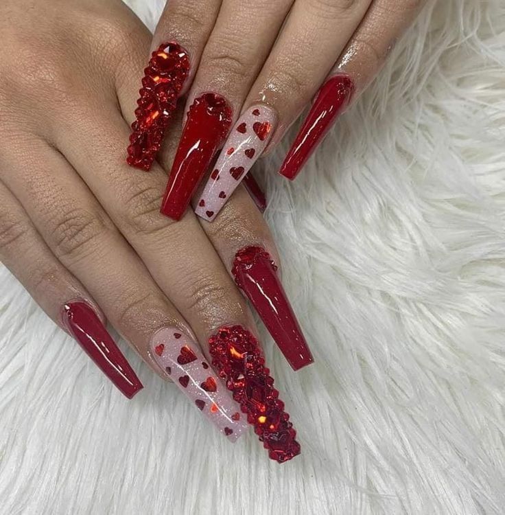 Long, coffin-shaped nails painted in deep red and nude with red heart decals, plus accent nails covered in red crystals.