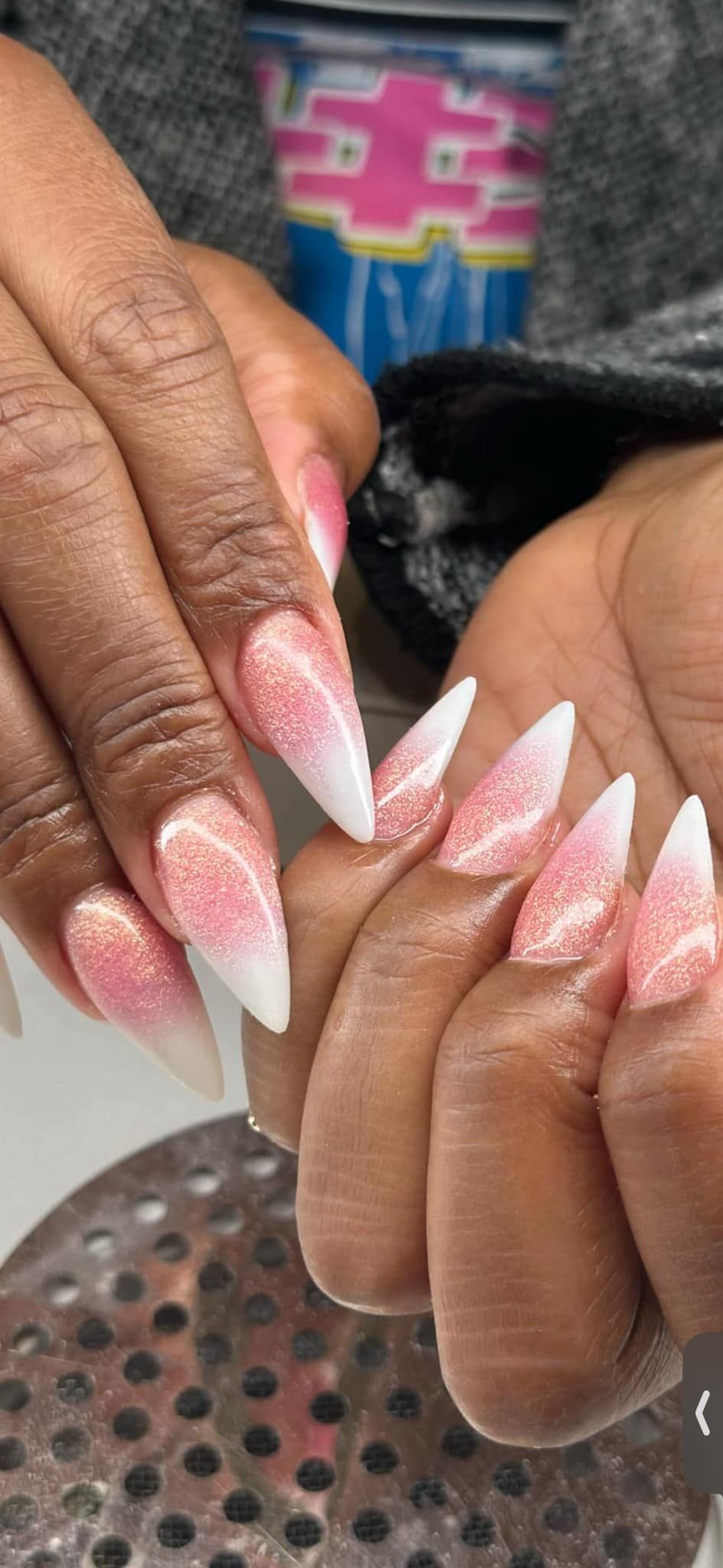 A close-up of hands showing stiletto-shaped acrylic nails with a pink and white glittery ombre design.