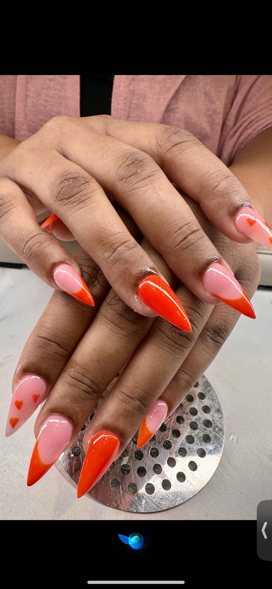 A person’s hands showcasing long, almond-shaped nails with a pink base and bright orange French tips and heart accents.