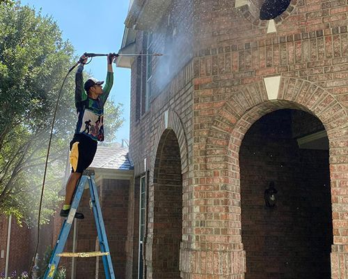 A man is standing on a ladder cleaning a brick building with a pressure washer.