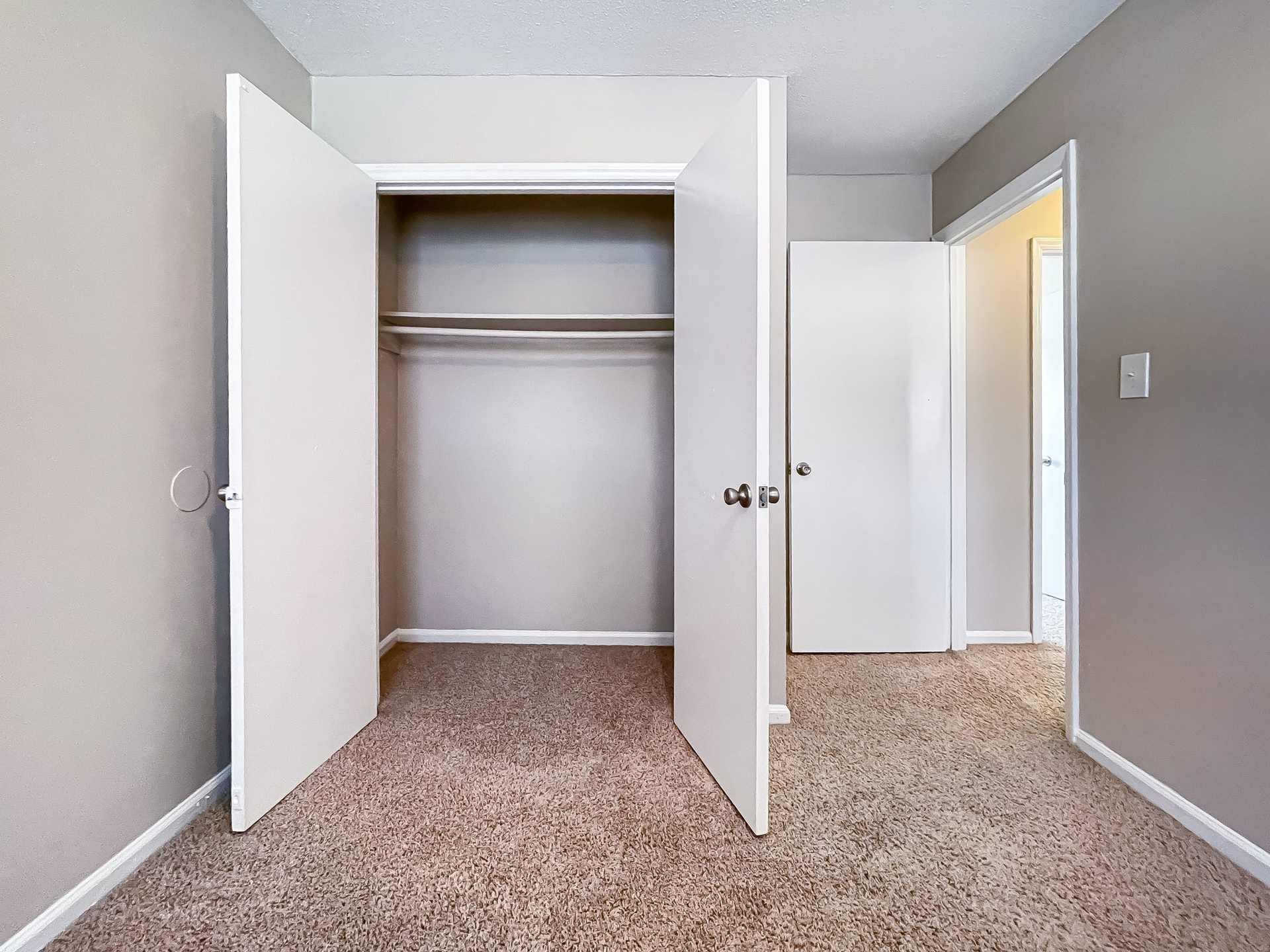 Open closet with double doors in a carpeted hallway, next to a doorway and gray walls.