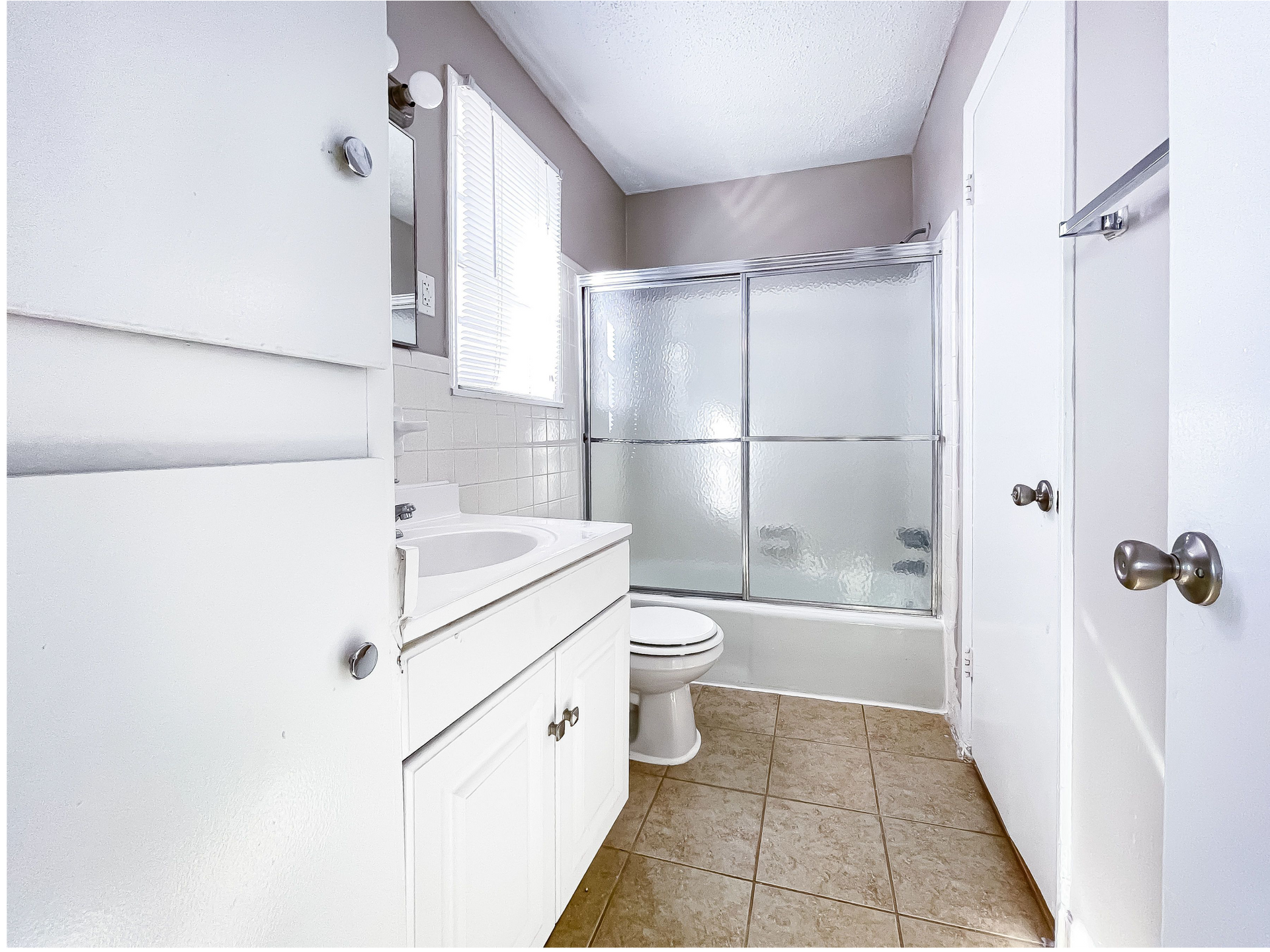 Small bathroom with white vanity, toilet, and frosted glass shower doors over tan tile floor.