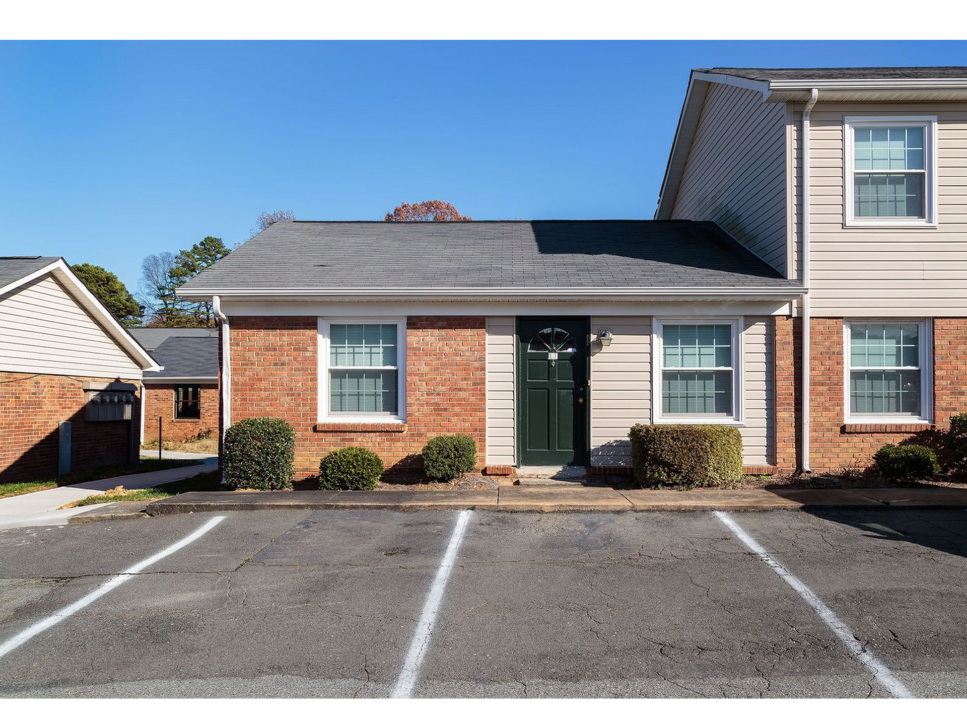Brick townhouse with green door and white-trimmed windows, viewed from a parking lot under a clear blue sky.