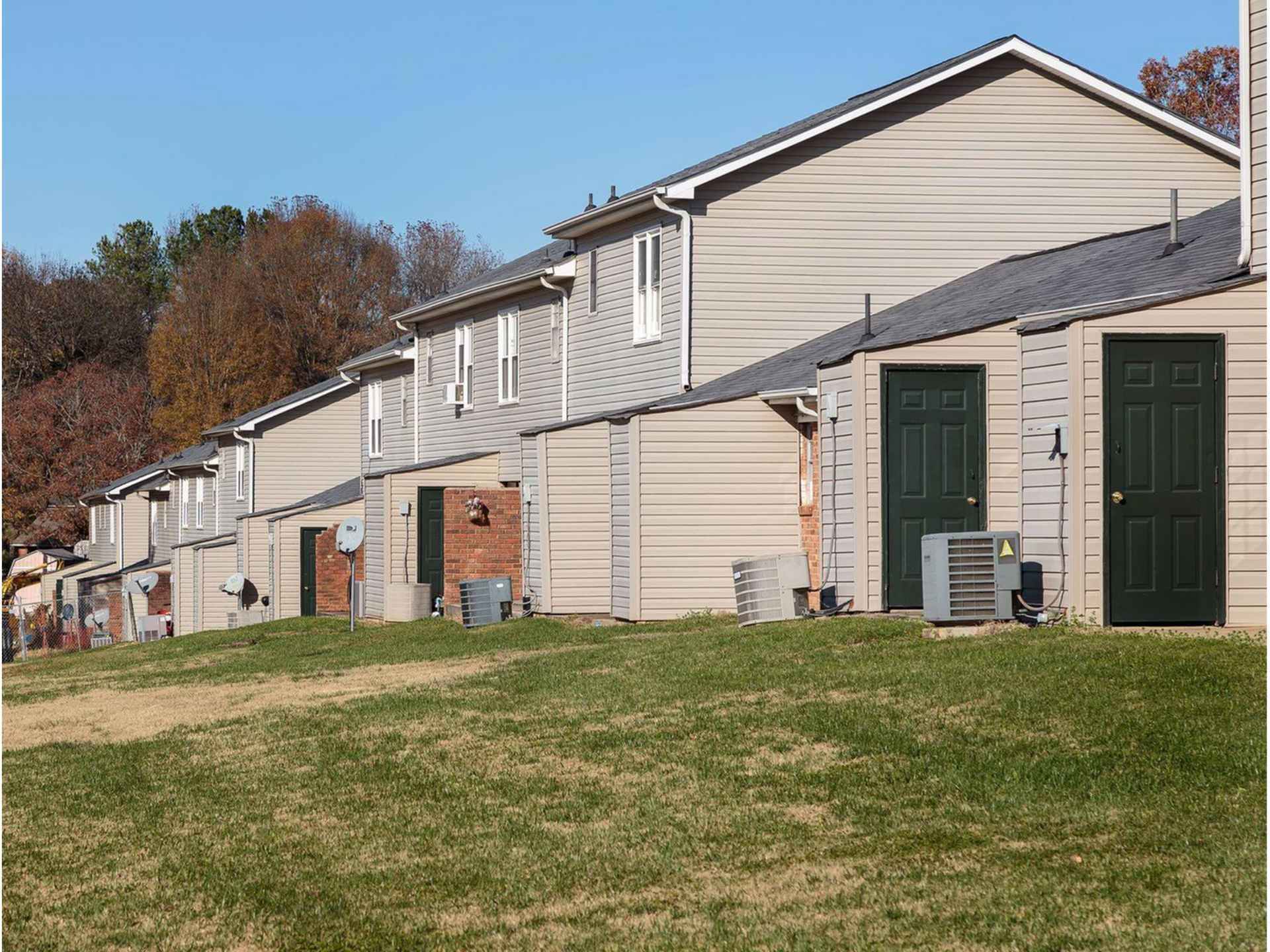 Row of beige townhouses with green doors along a grassy lawn under a clear sky