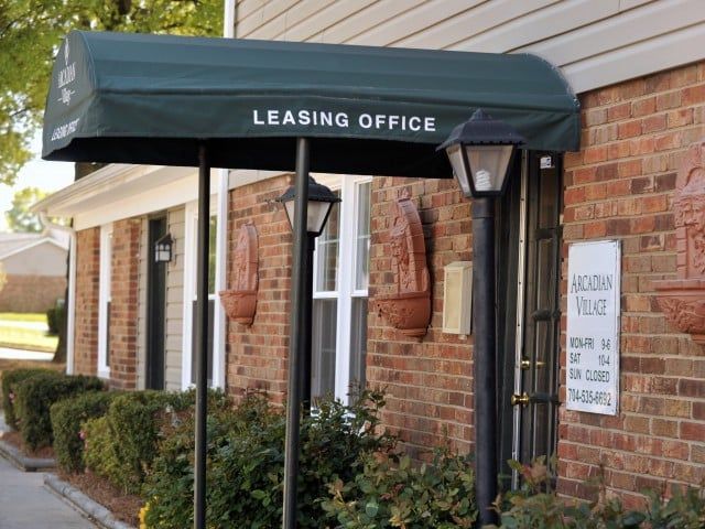 Brick apartment leasing office with green awning, windows, shrubs, and a sign by the entrance