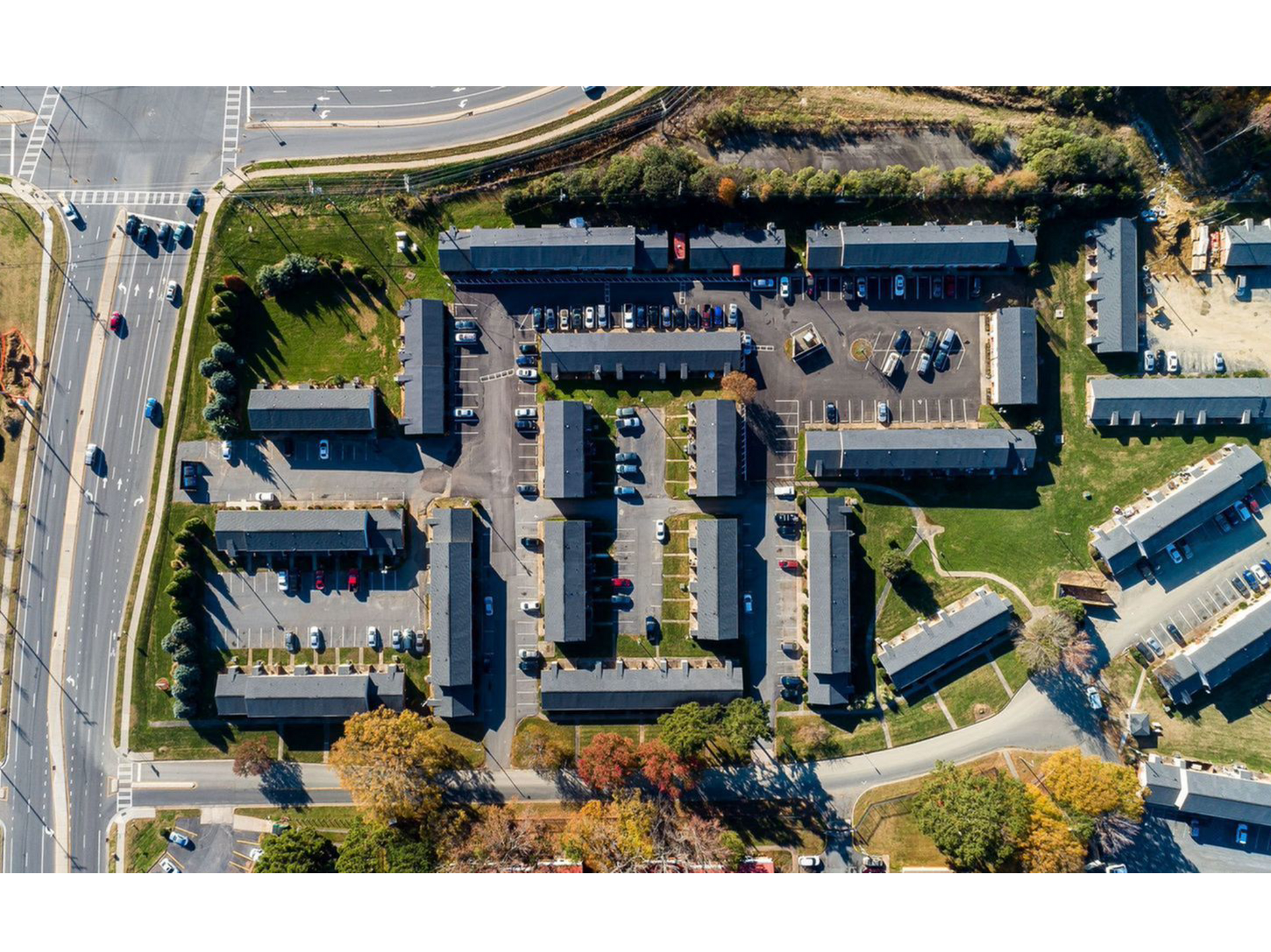 Aerial view of a residential apartment complex with parked cars, trees, and nearby roads.