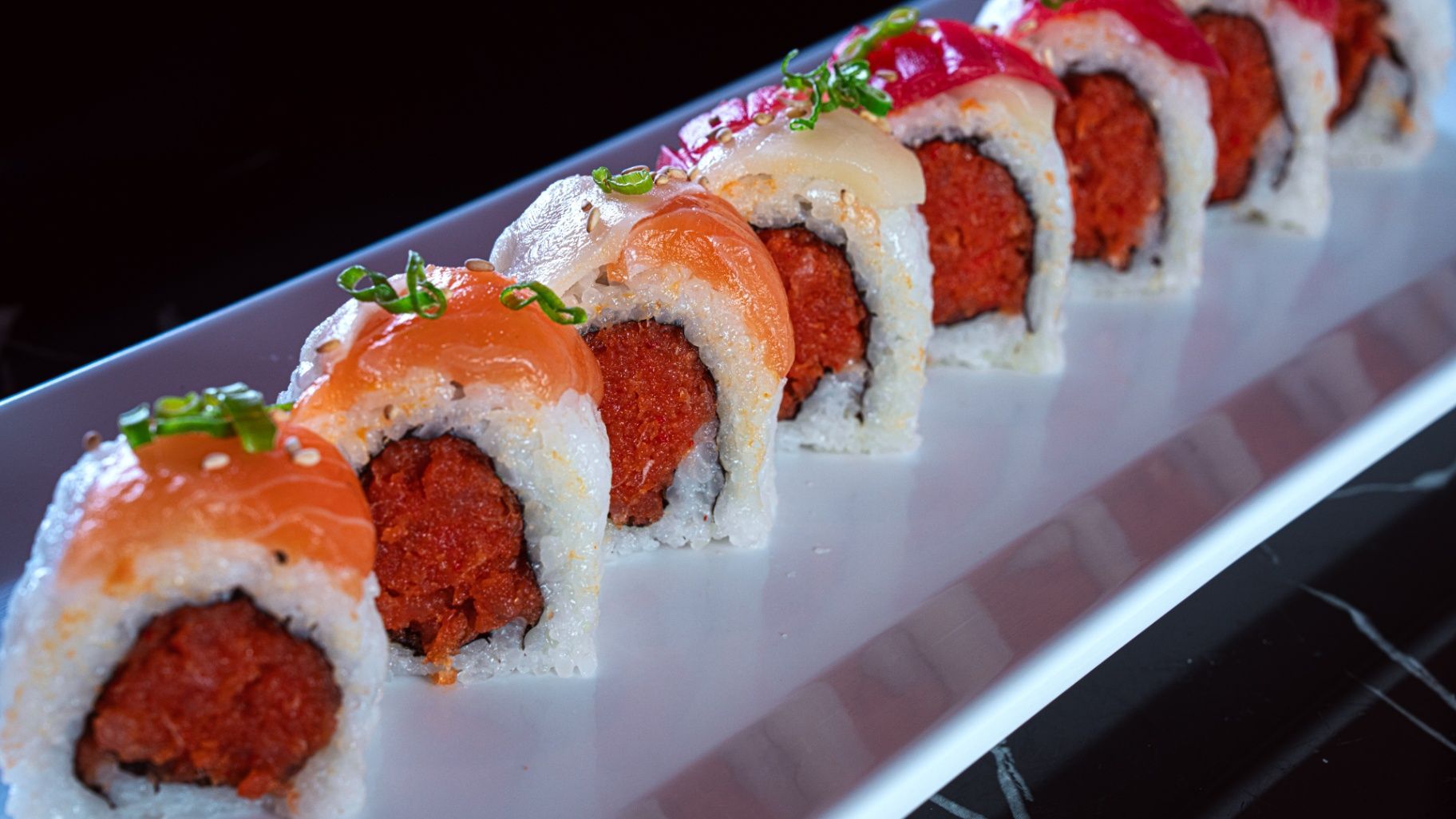 Sushi rolls with white rice and red filling, drizzled with sauce and topped with green garnish on a rectangular plate