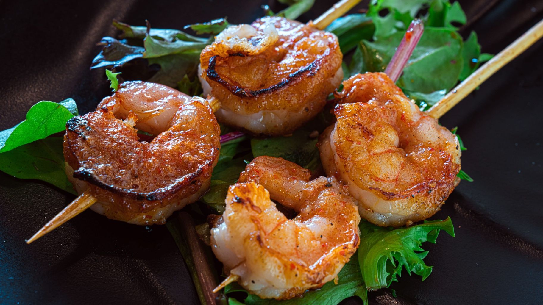 Grilled shrimp skewers on a dark plate with green garnish and glossy sauce