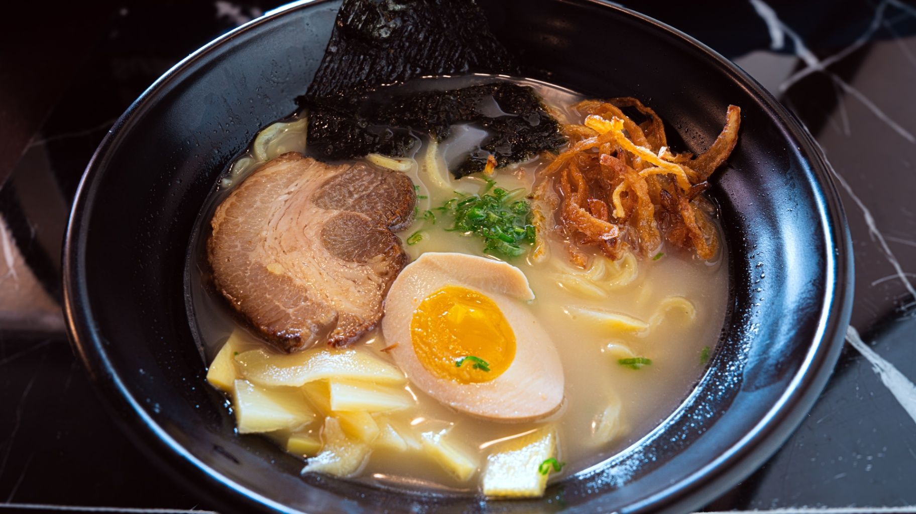 Bowl of ramen with sliced pork, soft-boiled egg, seaweed, and green onions in a dark bowl