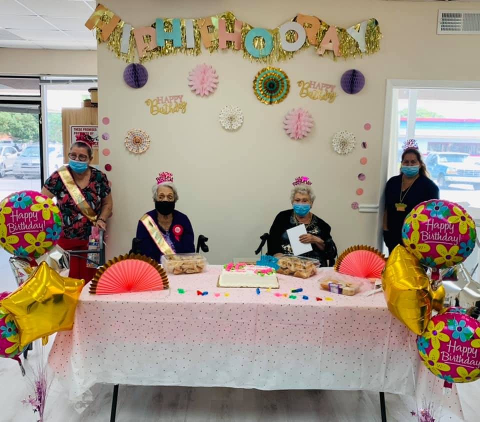 Birthday celebration: Two women seated at decorated table with staff. Balloons, cake, and banners in a room.