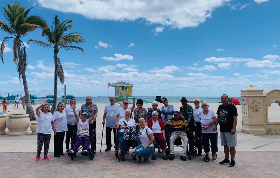 Group of people posing for a photo on a sunny beach, some using wheelchairs. Palm trees and lifeguard stand in the background