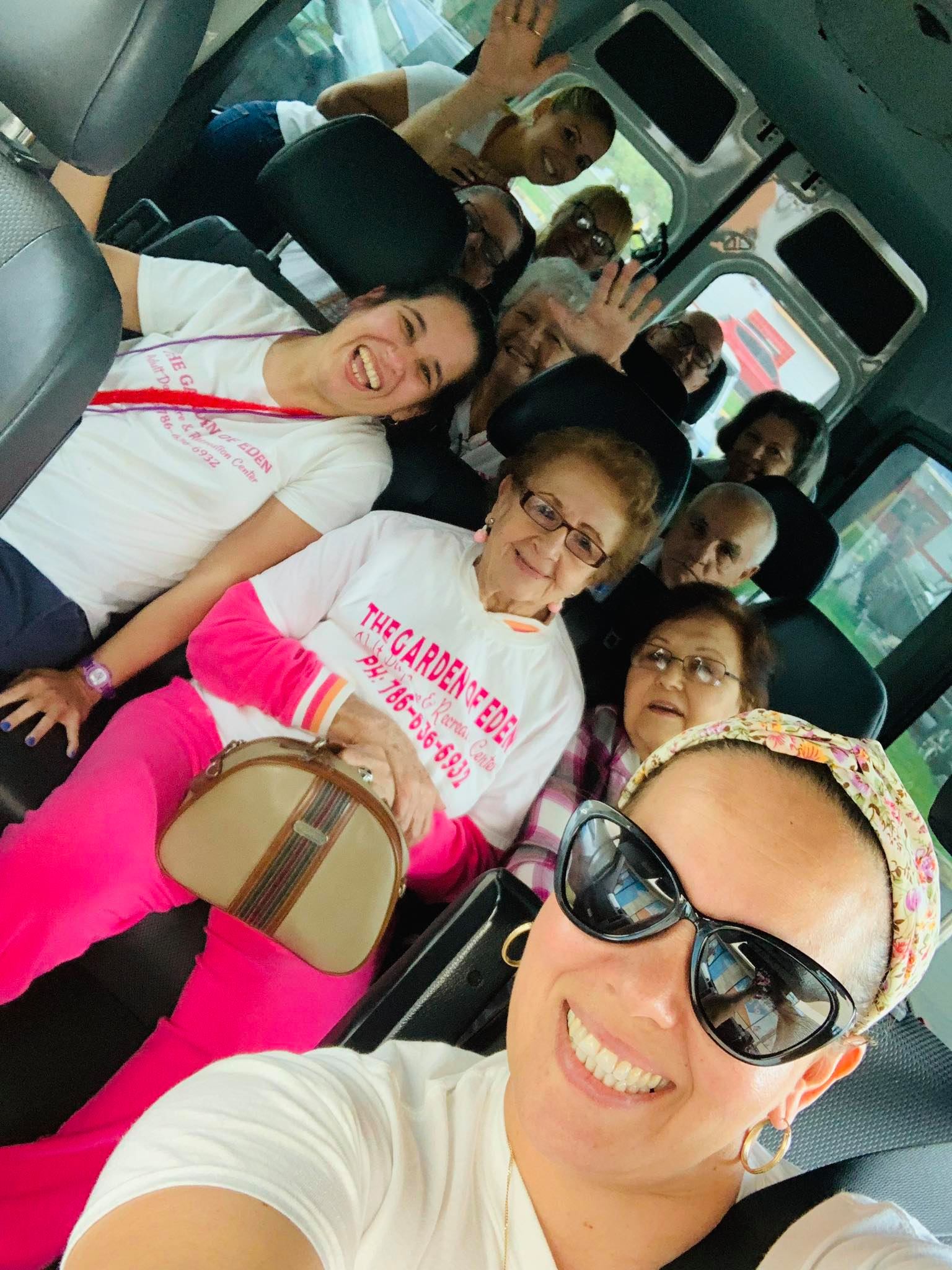 People smiling and waving inside a van. Some are wearing white shirts, one in pink pants and carrying a purse.