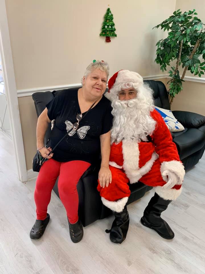 Woman in red pants sits with Santa Claus on a black couch, Christmas decor in background.