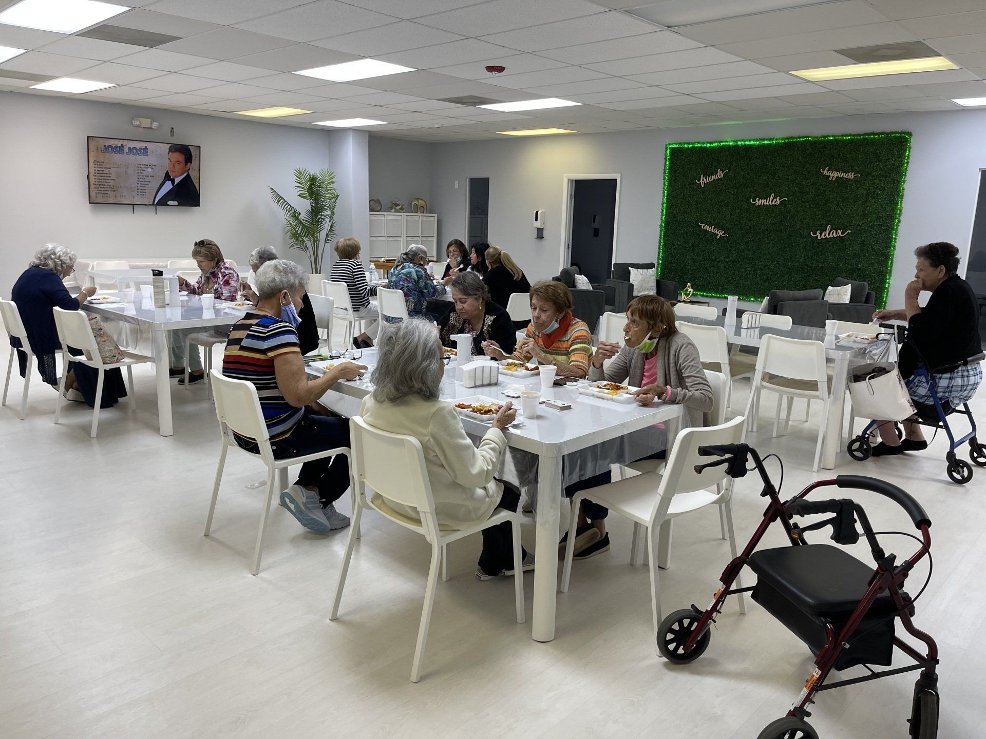 Senior citizens eat at tables in a brightly lit dining room. Some use walkers. A television is on the wall.