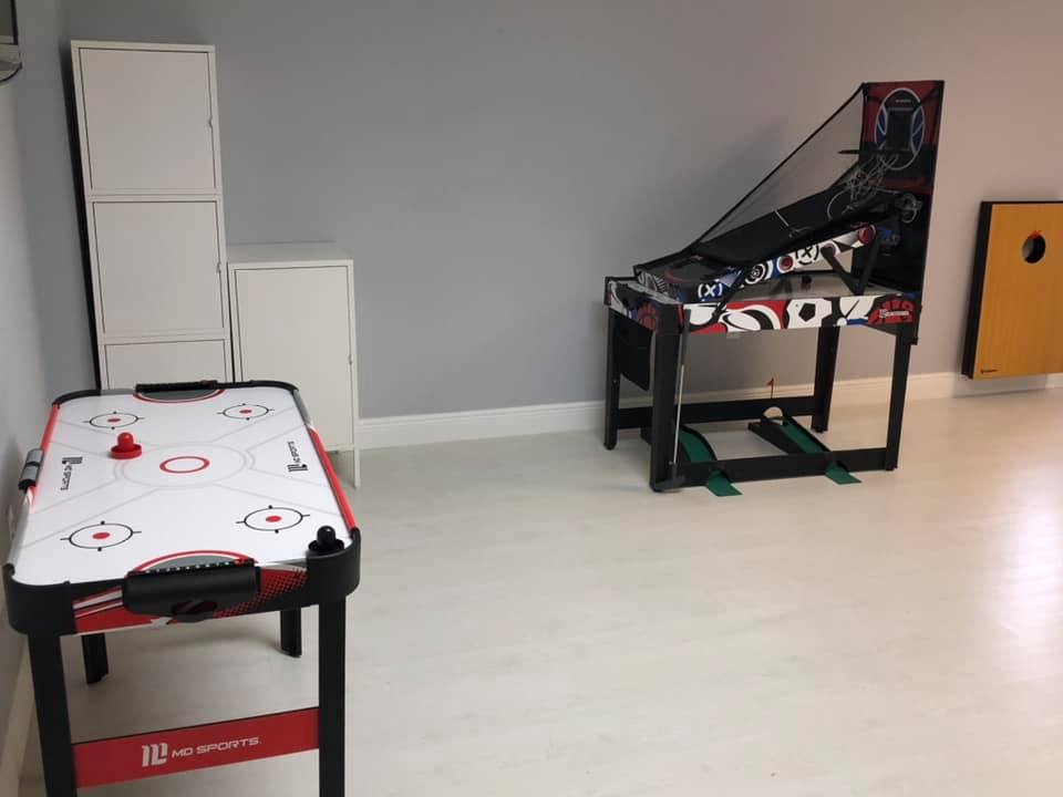 Air hockey table, skeeball game, and cornhole board in a rec room. White and red, on a white floor.