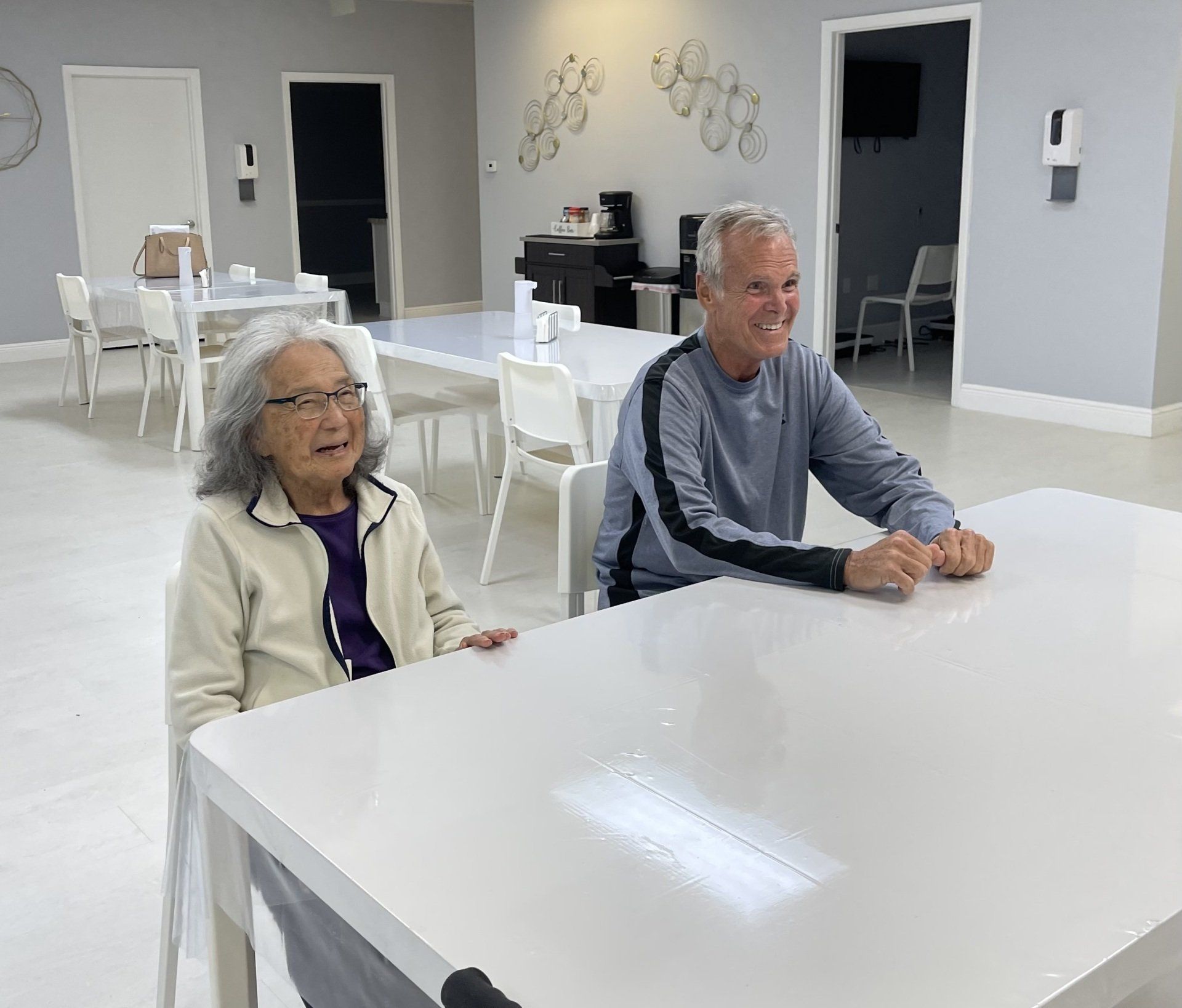 Two people sitting at a white table. The woman is smiling, the man looks happy. They are in a room with other tables.
