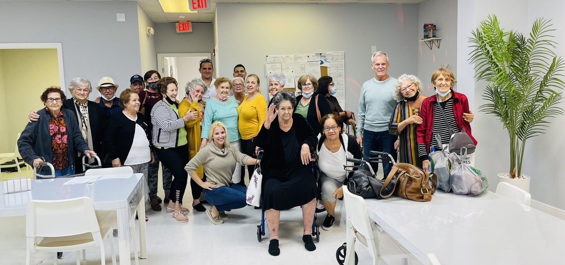 A group of people poses indoors, some smiling. There are tables, chairs, and plants.