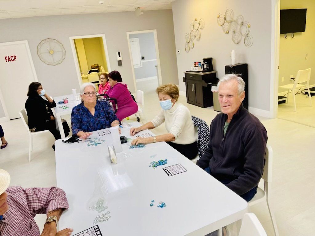 Seniors play a game at a table in a brightly lit room. Some wear masks.