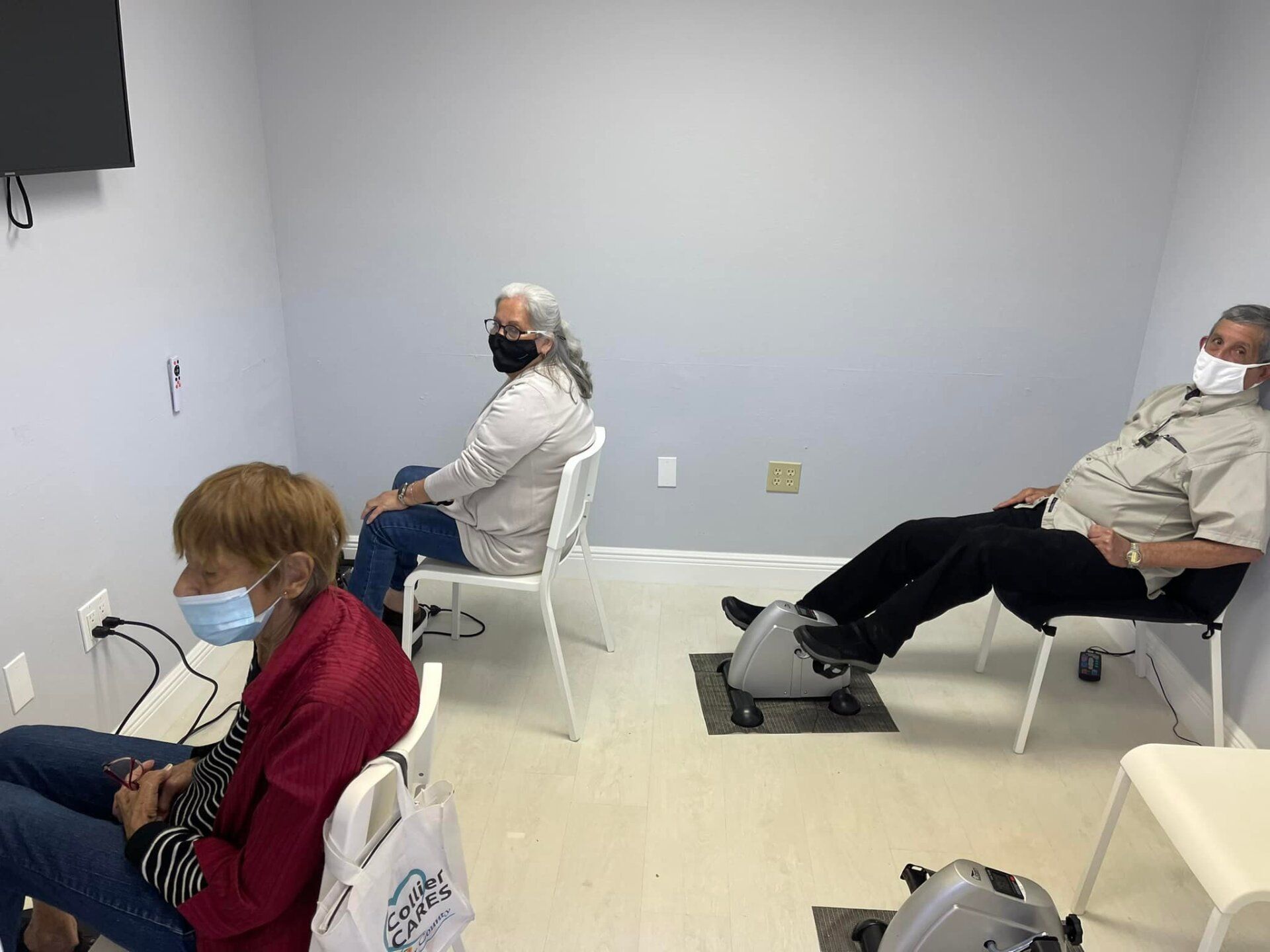 Three people in a room, wearing masks, seated and using small exercise machines, one pedaling. Gray walls.