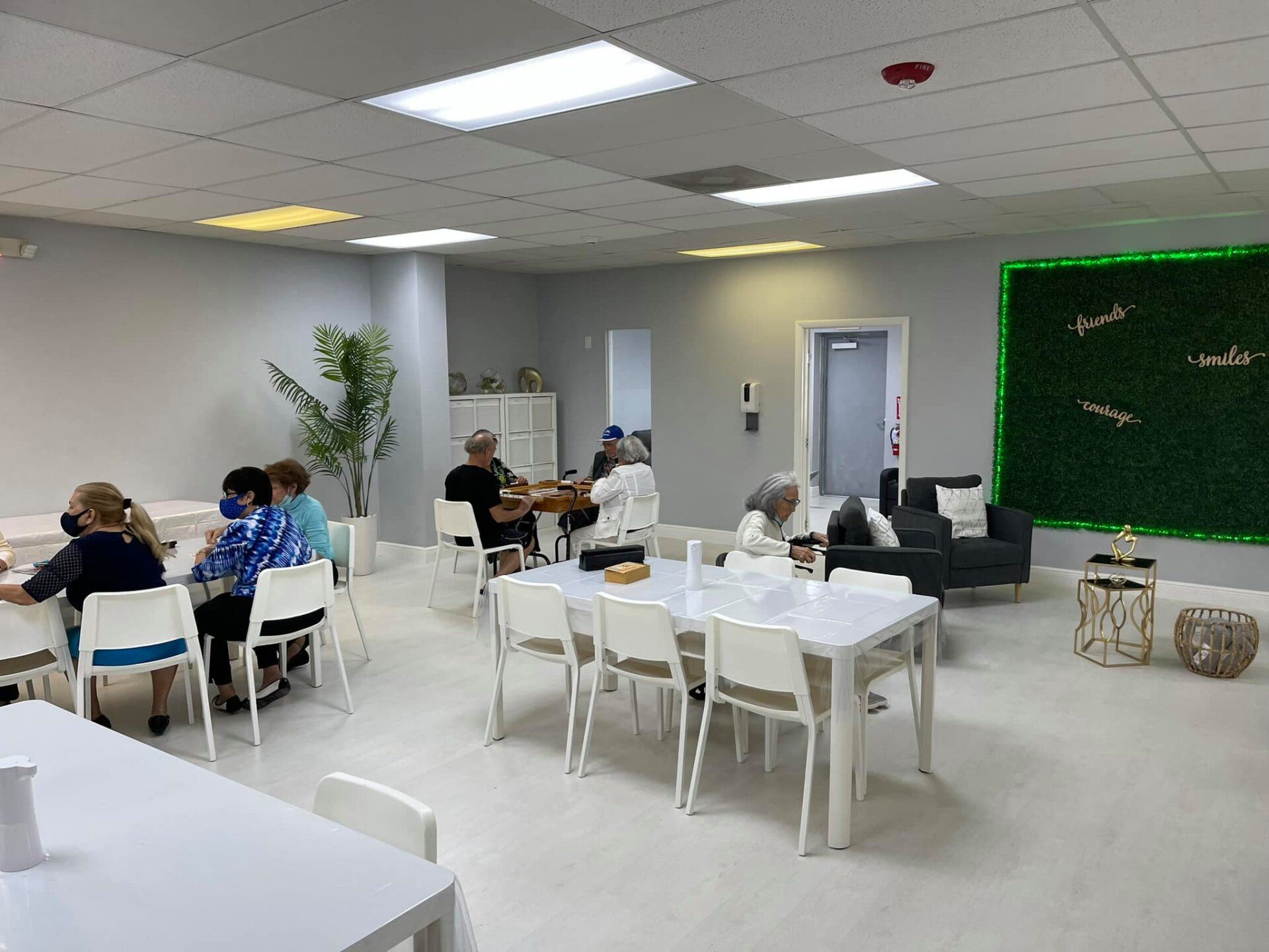 People seated at tables in a bright room, some wearing masks, a couch, and a green wall.
