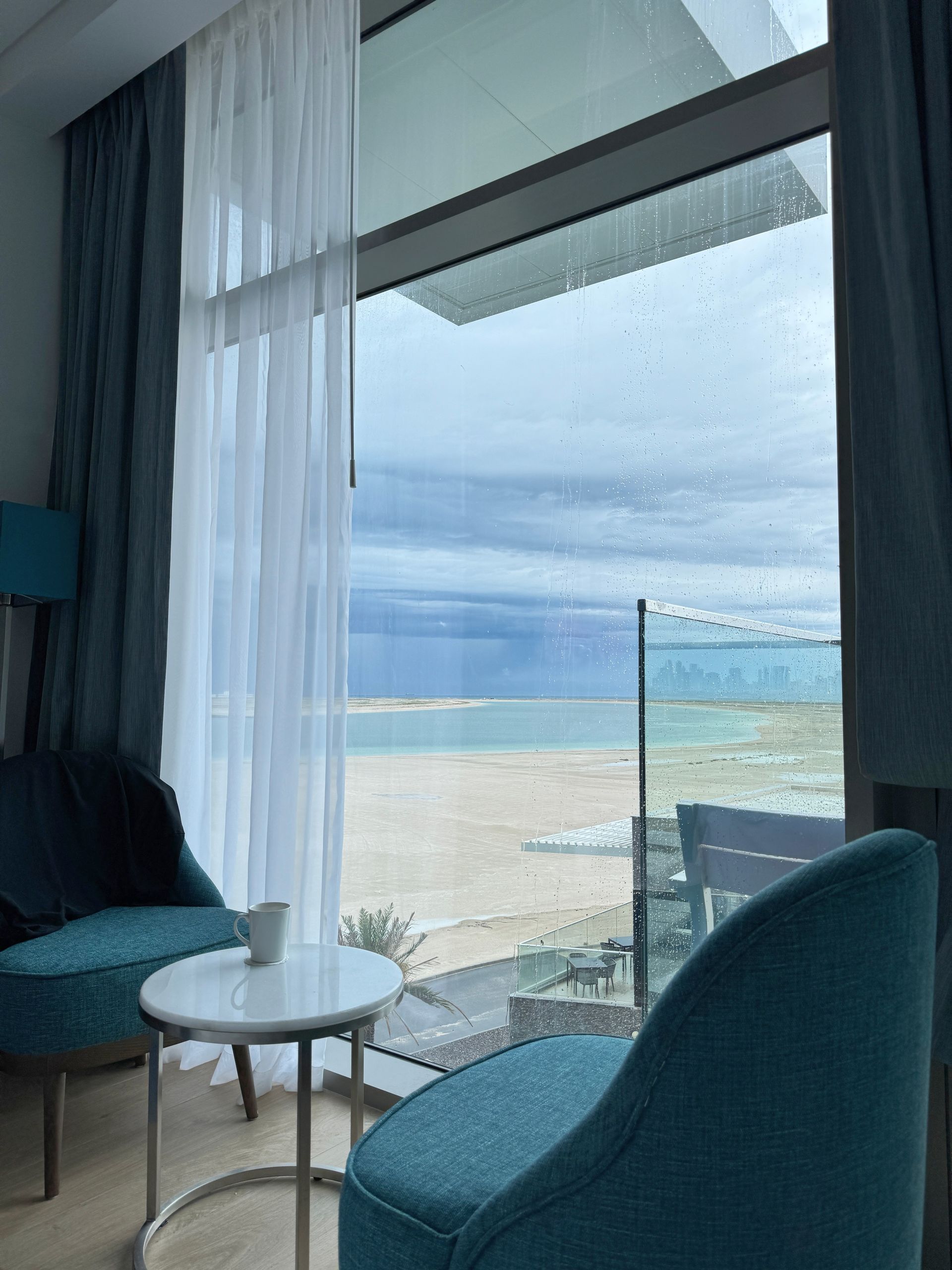 Cozy seaside room with teal chairs, small table, and a floor-to-ceiling window overlooking the beach and ocean
