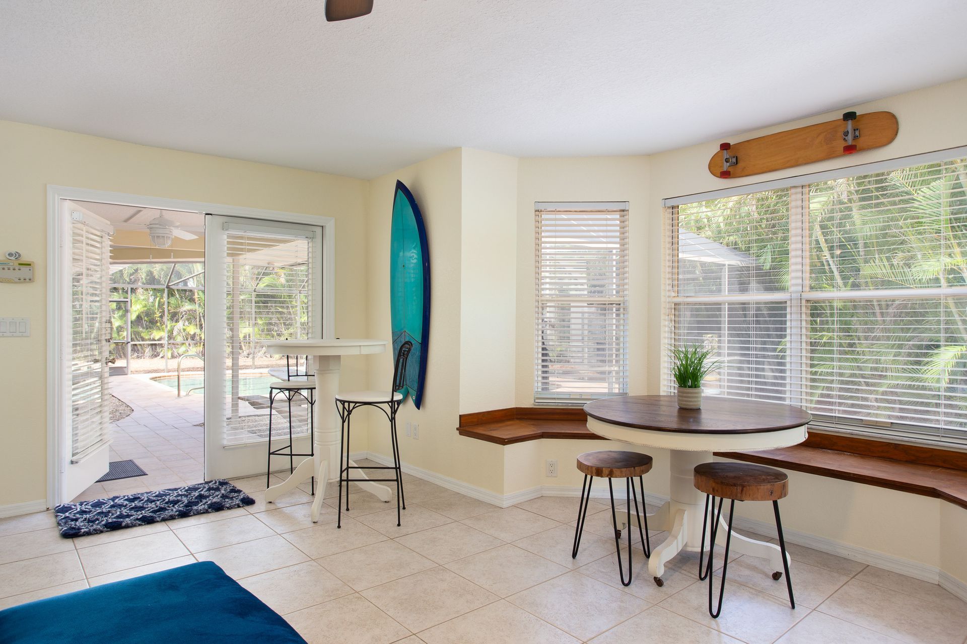 Bright dining nook with round table, stools, surfboard decor, and large windows opening to a patio.