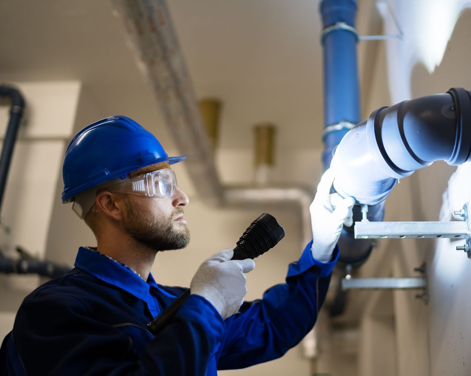 Un plombier en uniforme bleu, casque de chantier et lunettes de sécurité examine des tuyaux à l'aide d'une lampe torche dans un local technique.
